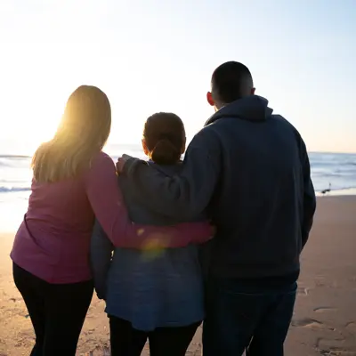 Three people standing together on the beach at sunset, embracing each other.