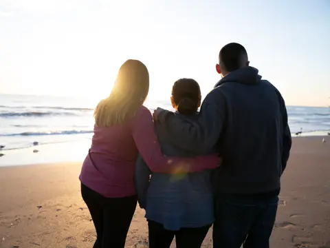 Three people standing together on the beach at sunset, embracing each other.