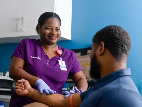 A smiling AdventHealth nurse in purple scrubs takes the blood pressure of a male patient.
