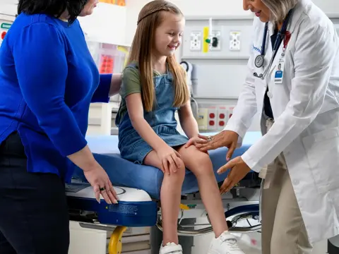A smiling young girl sits on a hospital bed while a doctor and nurse examine her leg.