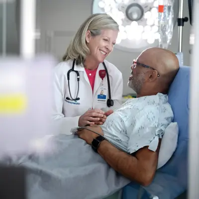 A smiling doctor in a white coat holds the hand of a patient lying in a hospital bed.