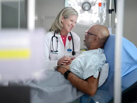 A smiling doctor in a white coat holds the hand of a patient lying in a hospital bed.