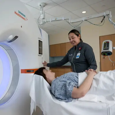 A smiling nurse assists a patient lying on a bed next to a large MRI machine.