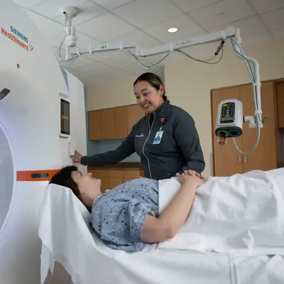 A smiling nurse assists a patient lying on a bed next to a large MRI machine.