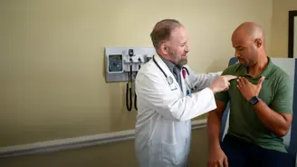 Doctor in white coat points to patient‚Äôs upper chest as patient touches chest during exam.
