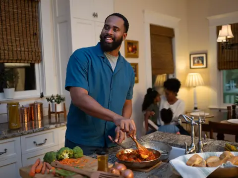 A man cooks in a kitchen with fresh vegetables and a family in the background.