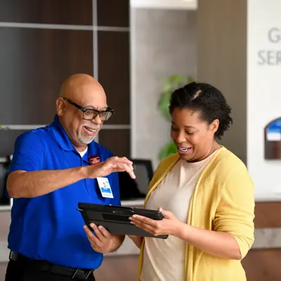 A smiling woman and a man in a blue shirt stand at a guest services desk, looking at a tablet.