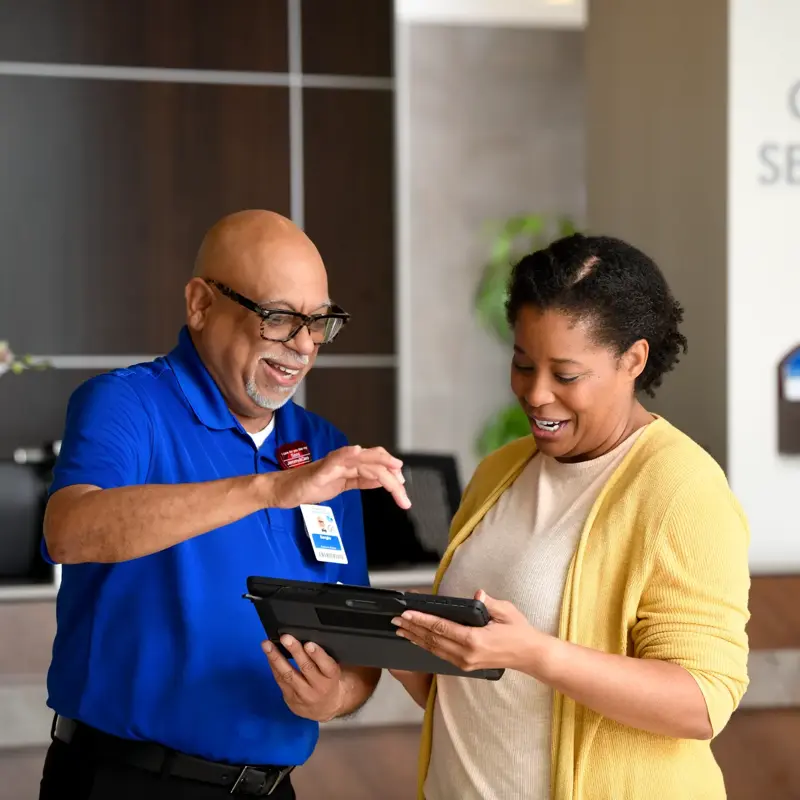A smiling woman and a man in a blue shirt stand at a guest services desk, looking at a tablet.
