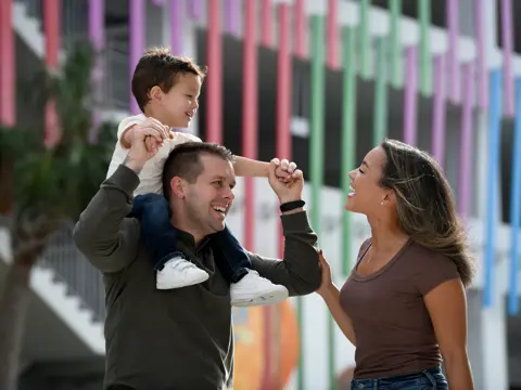 A father holds his son above his head while the mother looks on, all smiling.
