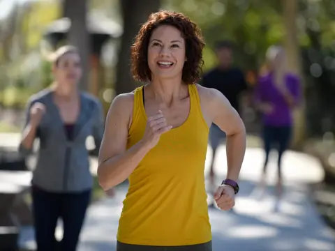 A woman in a yellow tank top is jogging on a path with other people in the background.