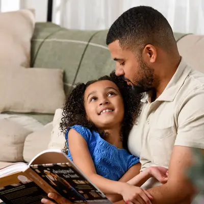 Father and young daughter sitting together on a couch reading a book in a bright living room