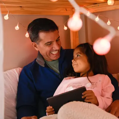 A father and daughter are smiling while reading a book on a tablet in a cozy bedroom.