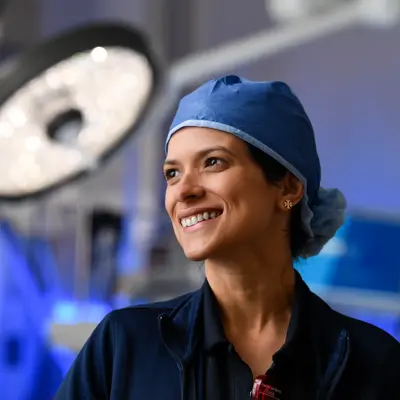 Smiling female surgeon in blue surgical cap and scrubs looking upward in an operating room with surgical lights visible in the background
