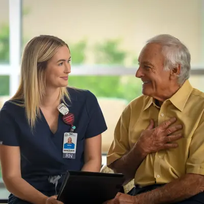 A nurse in blue scrubs sits with an elderly man, both smiling and engaged in conversation.