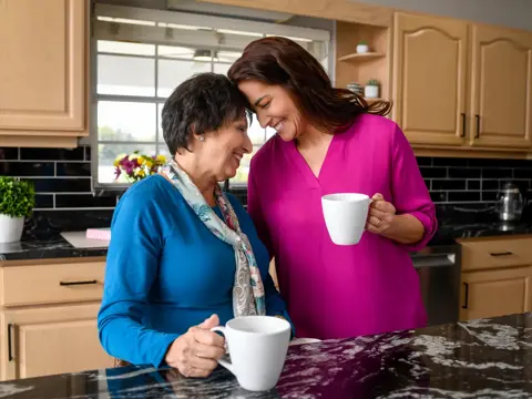Two women in a kitchen, smiling and holding coffee cups, sharing a moment of connection.