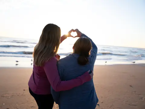 Two women embrace on the beach at sunset, forming a heart shape with their hands.