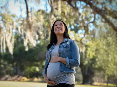 Pregnant woman in denim jacket and gray top smiling while looking up outdoors in a park setting