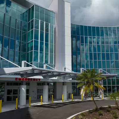 Modern hospital building with glass facade, emergency entrance, and palm trees.