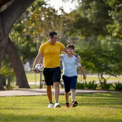 Father and son walking in park, holding hands and soccer ball.