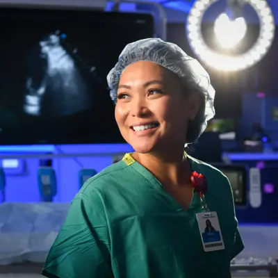 Smiling female healthcare worker in scrubs and hairnet in a hospital room with medical equipment.