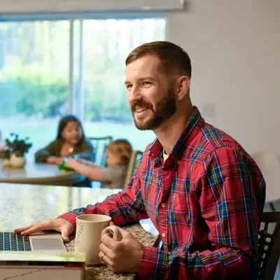 A man in a plaid shirt works on his laptop at home with his family in the background.