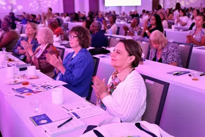 Attendees applauding during a women’s health event in a conference ballroom illuminated with pink and purple lighting.