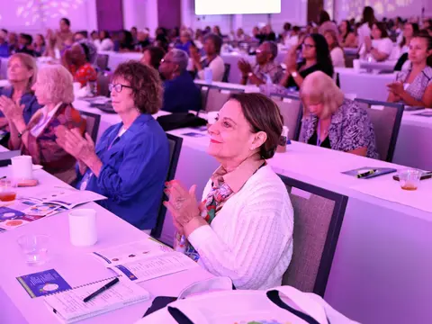 Attendees applauding during a women’s health event in a conference ballroom illuminated with pink and purple lighting.