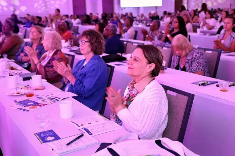 Attendees applauding during a women’s health event in a conference ballroom illuminated with pink and purple lighting.