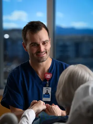 Healthcare worker in blue scrubs smiles while holding an older patient’s hands beside a hospital bed.