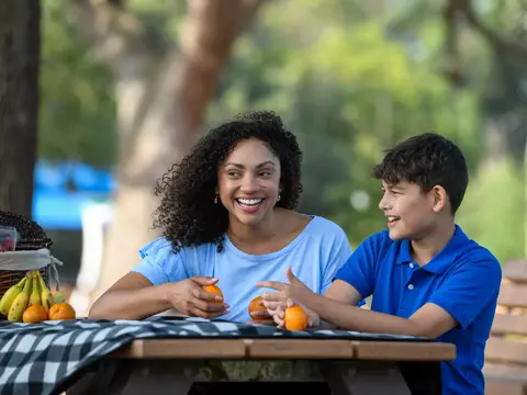 A smiling woman and boy sit at a picnic table with fruit, enjoying a sunny day.