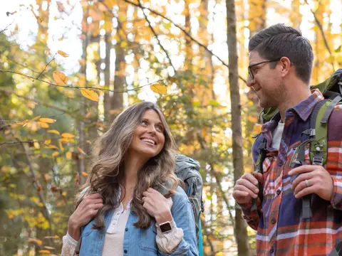 A smiling woman and man hiking in the woods, both wearing backpacks and enjoying the outdoors.