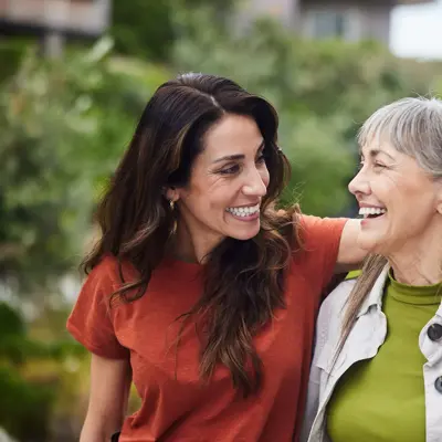 Two women walking outdoors with arms around each other, smiling and looking at one another.