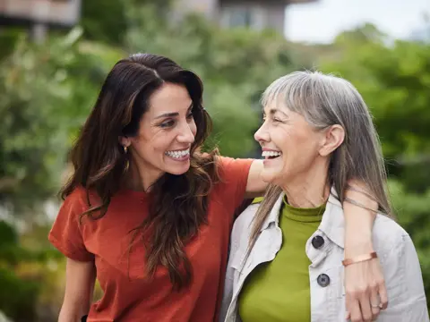 Two women walking outdoors with arms around each other, smiling and looking at one another.