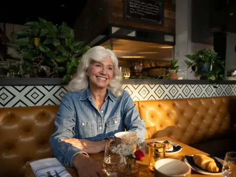 A smiling woman with white hair sits at a table in a restaurant, surrounded by plants and decor.