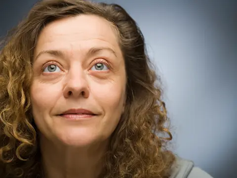 Woman with curly brown hair looks slightly upward against a gradient blue-gray background.