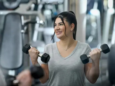 A smiling woman in a gray shirt holds dumbbells in a gym.