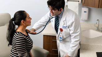 Doctor in white coat using a stethoscope to examine a seated female patient in a medical exam room