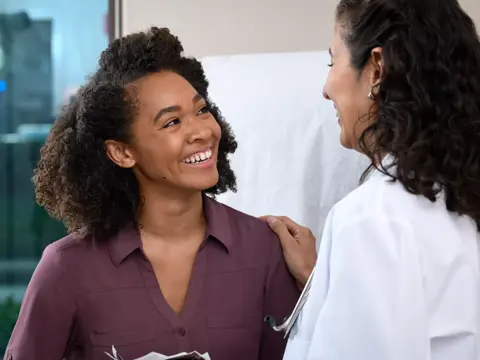 A smiling woman in a maroon shirt is being examined by a doctor in a white coat.