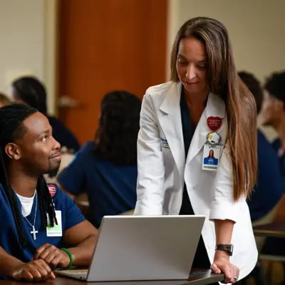 Instructor in white coat assisting a student in navy scrubs at a laptop in a classroom filled with other students