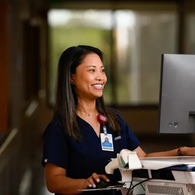 Smiling nurse in navy scrubs sitting at a computer workstation in a health care facility