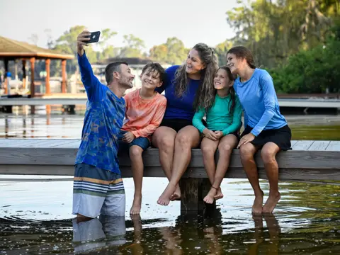 Family of four smiling and taking a selfie on a wooden dock by the water.
