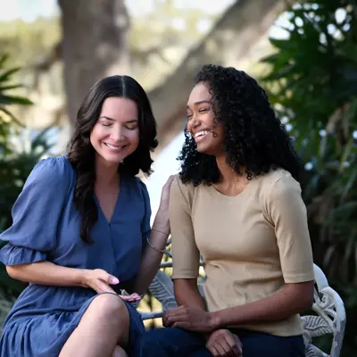 Two women sitting together on a white bench outdoors, smiling and looking at a smartphone in a lush garden setting