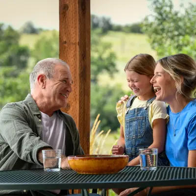 A grandfather, mother, and daughter laugh together at an outdoor table with a bowl of fruit.