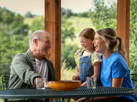 A grandfather, mother, and daughter laugh together at an outdoor table with a bowl of fruit.