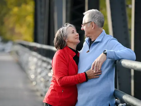 Elderly couple standing close together on a bridge, smiling and looking at each other.