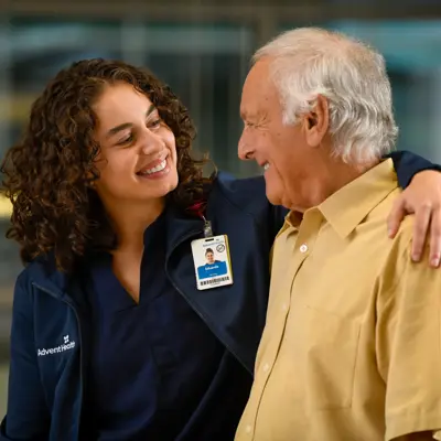 A smiling nurse with curly hair embraces an elderly man, both wearing AdventHealth badges.