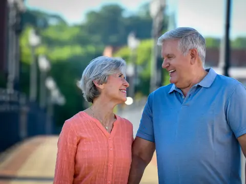Smiling senior couple in casual clothing standing together outdoors on a sunny day with greenery and a walkway in the background