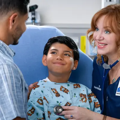 A smiling nurse uses a stethoscope to listen to a young boy's heart while his father watches.