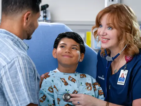 A smiling nurse uses a stethoscope to listen to a young boy's heart while his father watches.