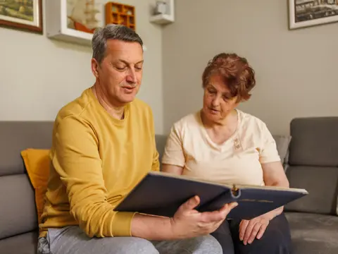 Two adults sit on a couch, looking at an open photo album together in a living room.